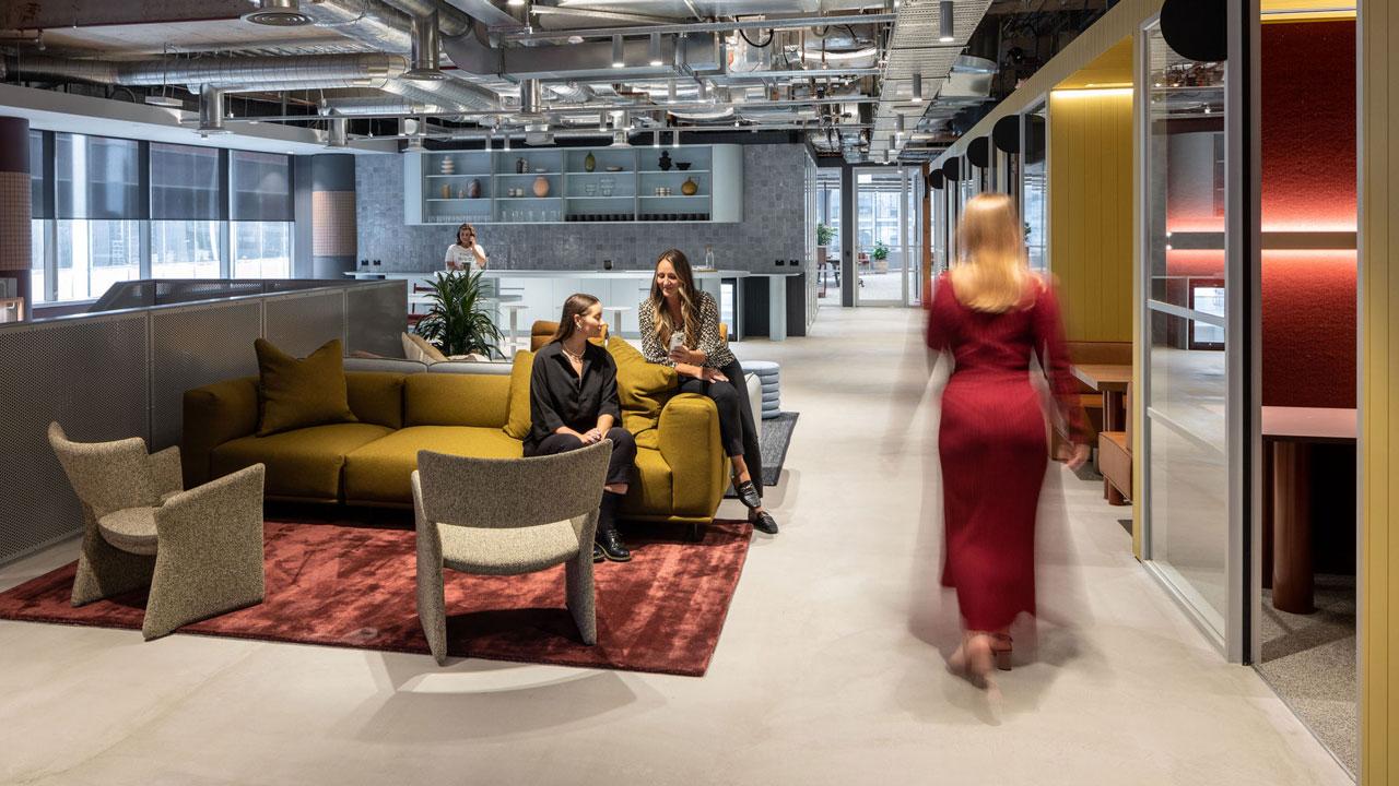 A woman walks through an open space with a mix of seating types, colorful accent walls and furniture, and exposed ductwork